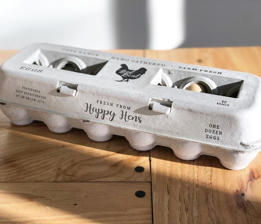 A Henlay Laid Local Egg Carton by My Pet Chicken, featuring an adorable printed vintage design, sits on a wooden table in bright natural light. The light gray recycled pulp fiber carton holds one dozen eggs.