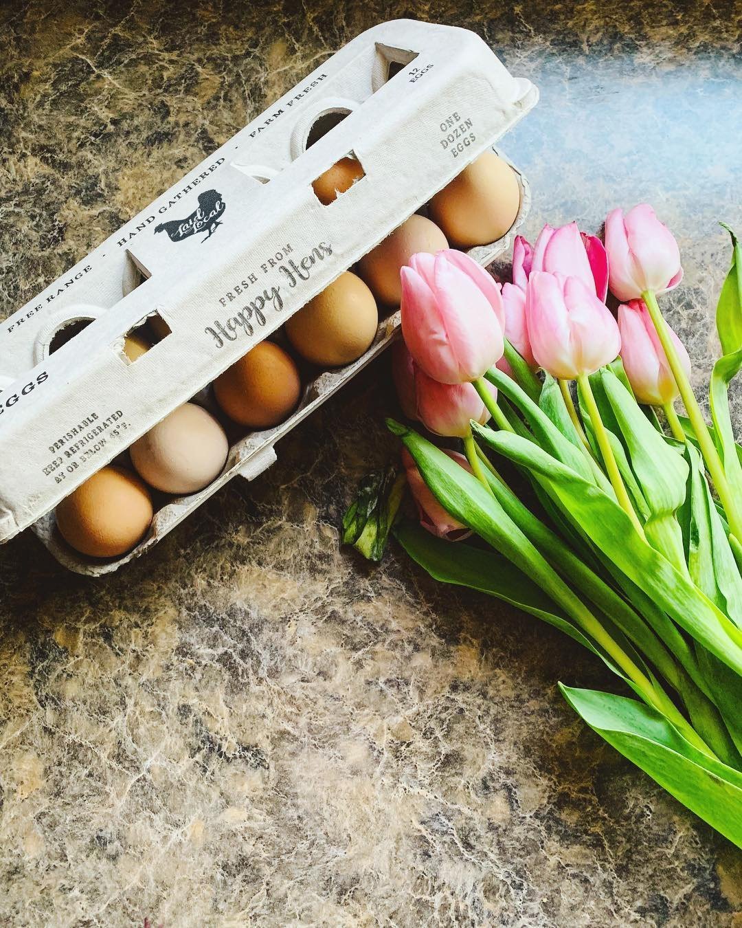 A carton of My Pet Chicken Henlay Laid Local Egg Cartons with an adorable vintage design rests on a marble countertop beside pink tulips with green stems.