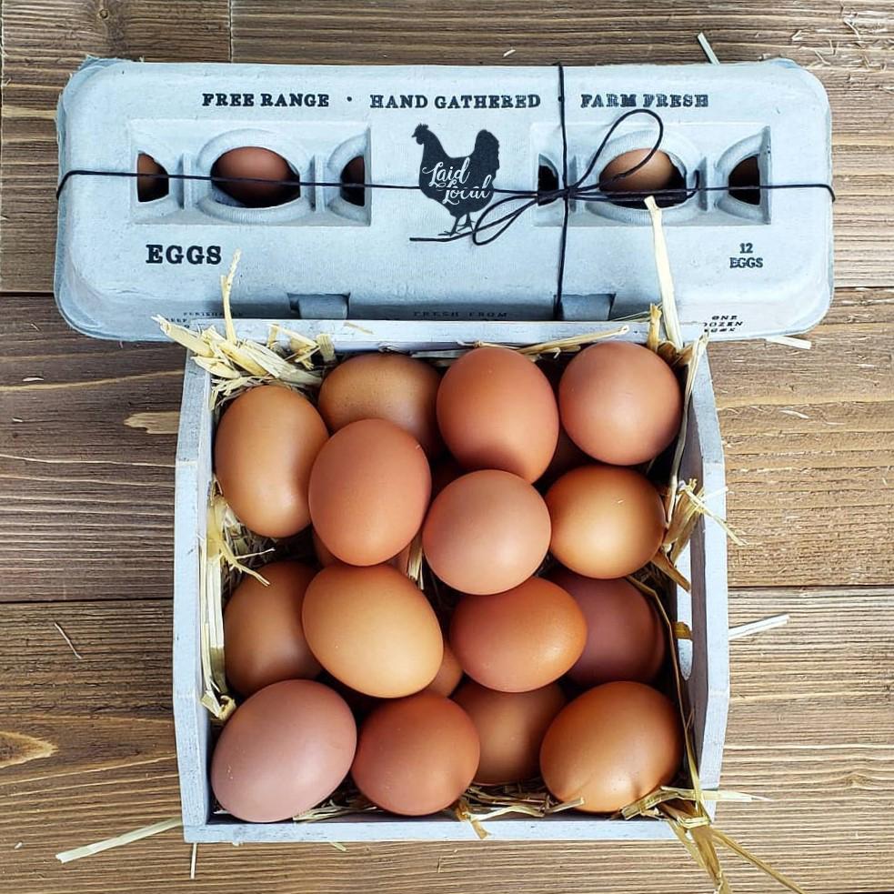 A carton of Henlay Laid Local Egg Cartons by My Pet Chicken, featuring an adorable vintage design, is open next to a straw-filled basket of brown eggs on wood—available in packs of 25, 90, or 250.