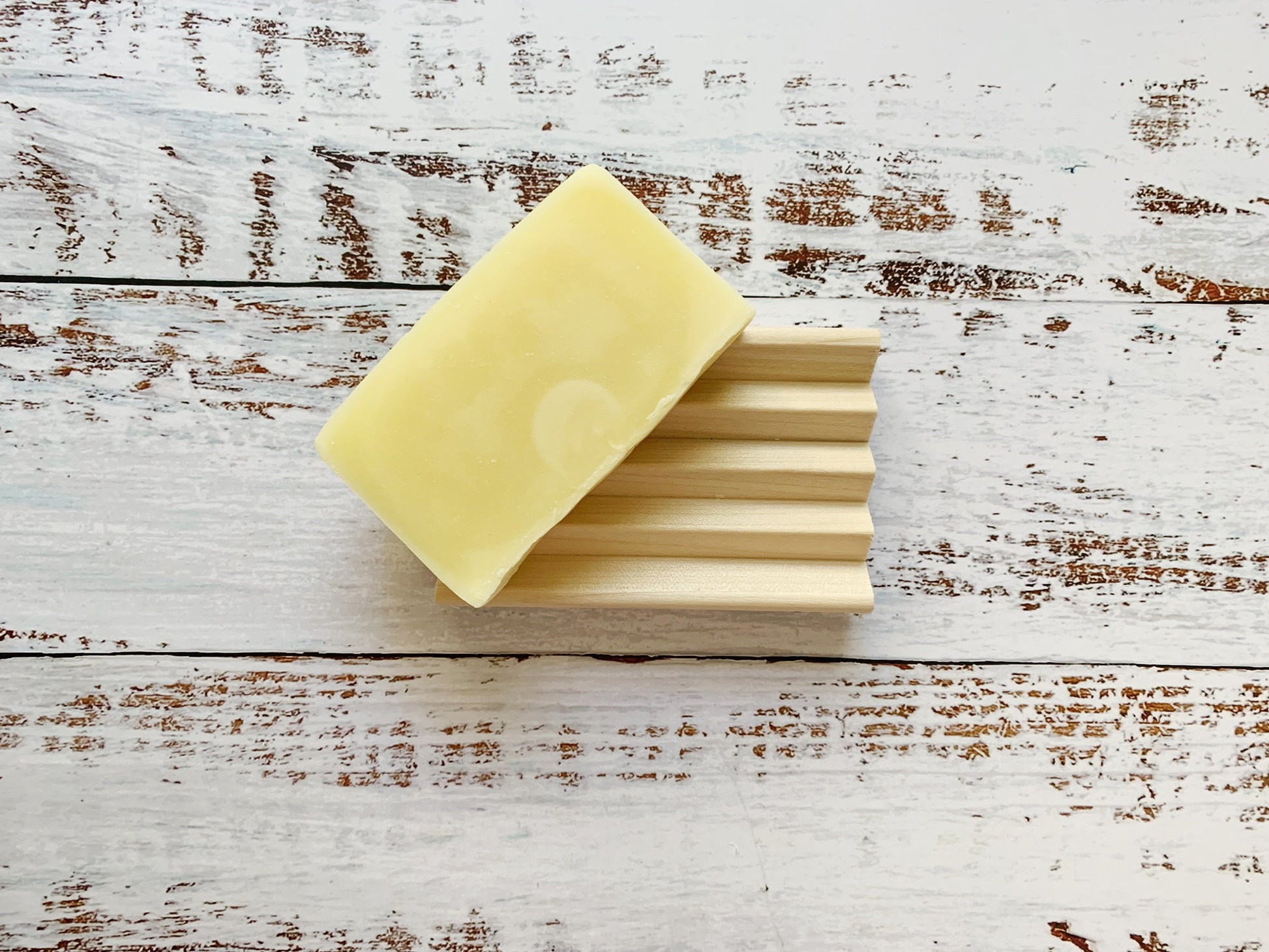 A pale yellow soap bar sits on a deeply grooved Wooden Soap Dish from Dot and Army (set of two), displayed atop a rustic white wooden surface with visible brown grain and weathered texture.
