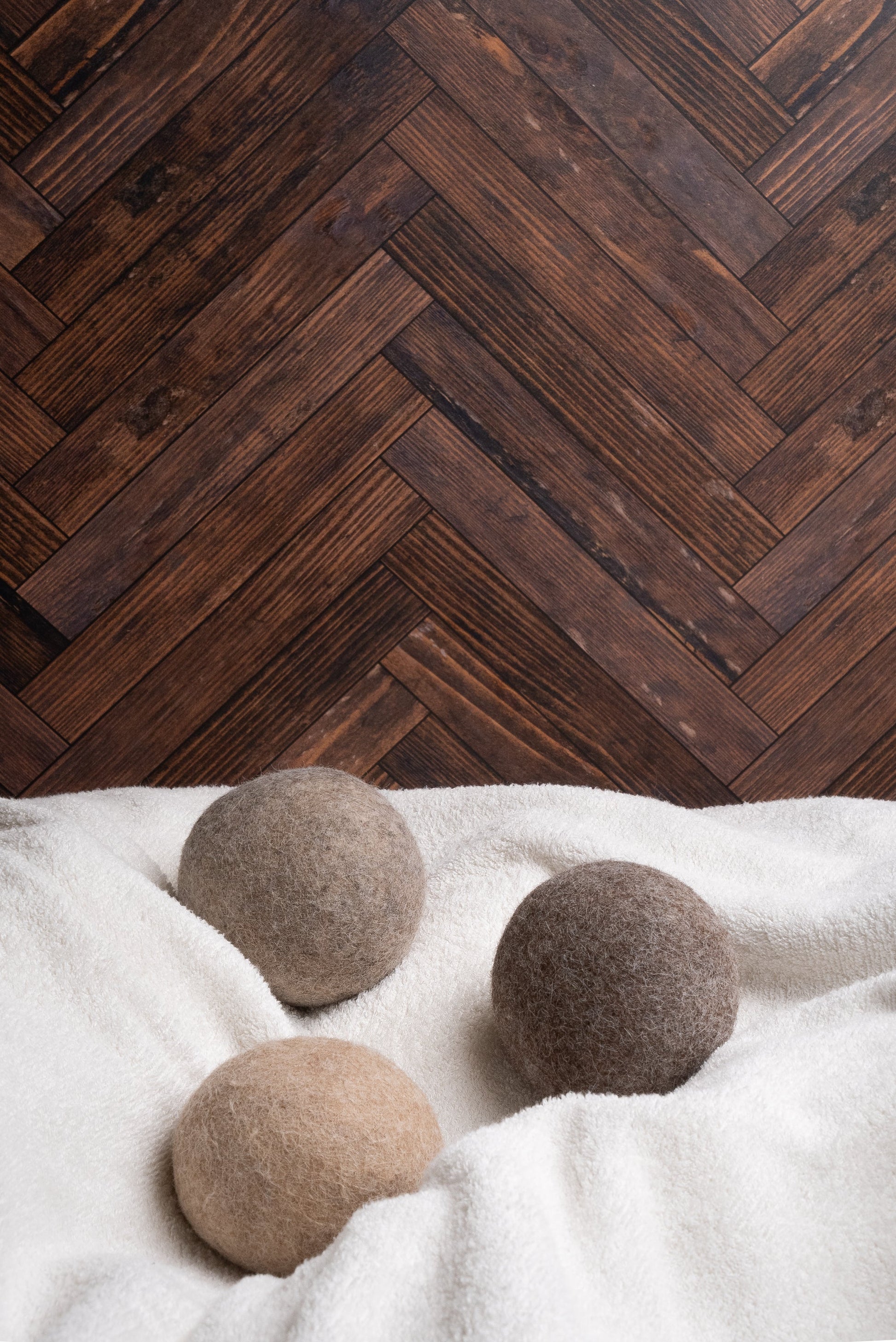 Rustic Strength’s Alpaca Dryer Balls (3 pack) in beige, brown, and gray rest on a white towel, providing a natural wool alternative with static reduction. The scene is set against a dark wooden herringbone-patterned floor.