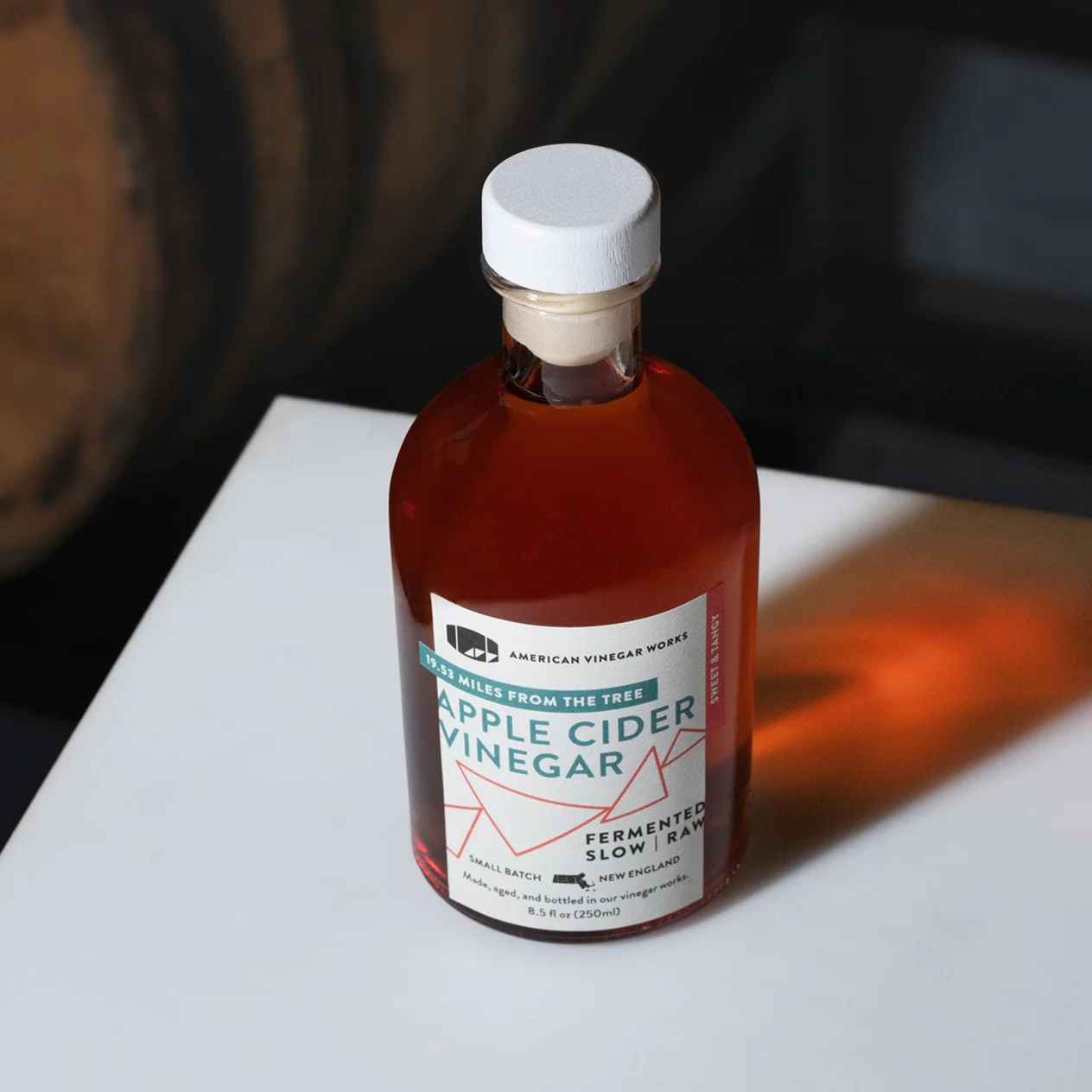 A bottle of American Vinegar Works Apple Cider Vinegar with a natural lifestyle scene featuring apples and a rustic background.