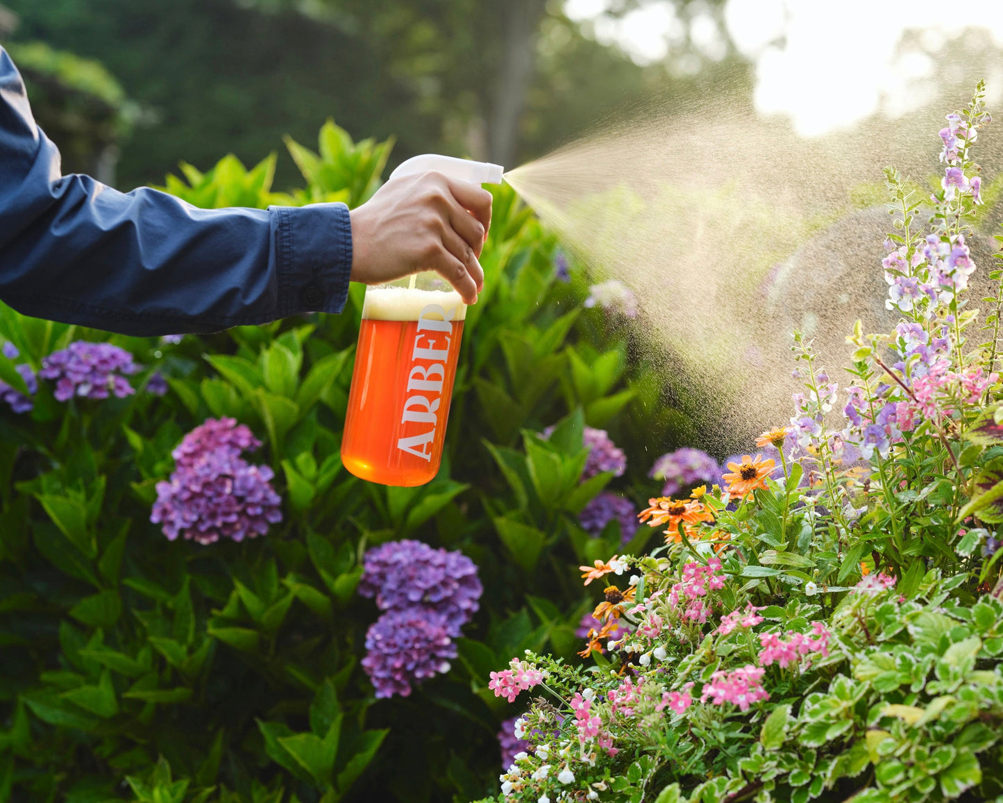 Using an Arber Spray Bottle, a person mists blooming garden flowers, supporting plant health as sunlight catches the fine spray and bright blossoms in the background.