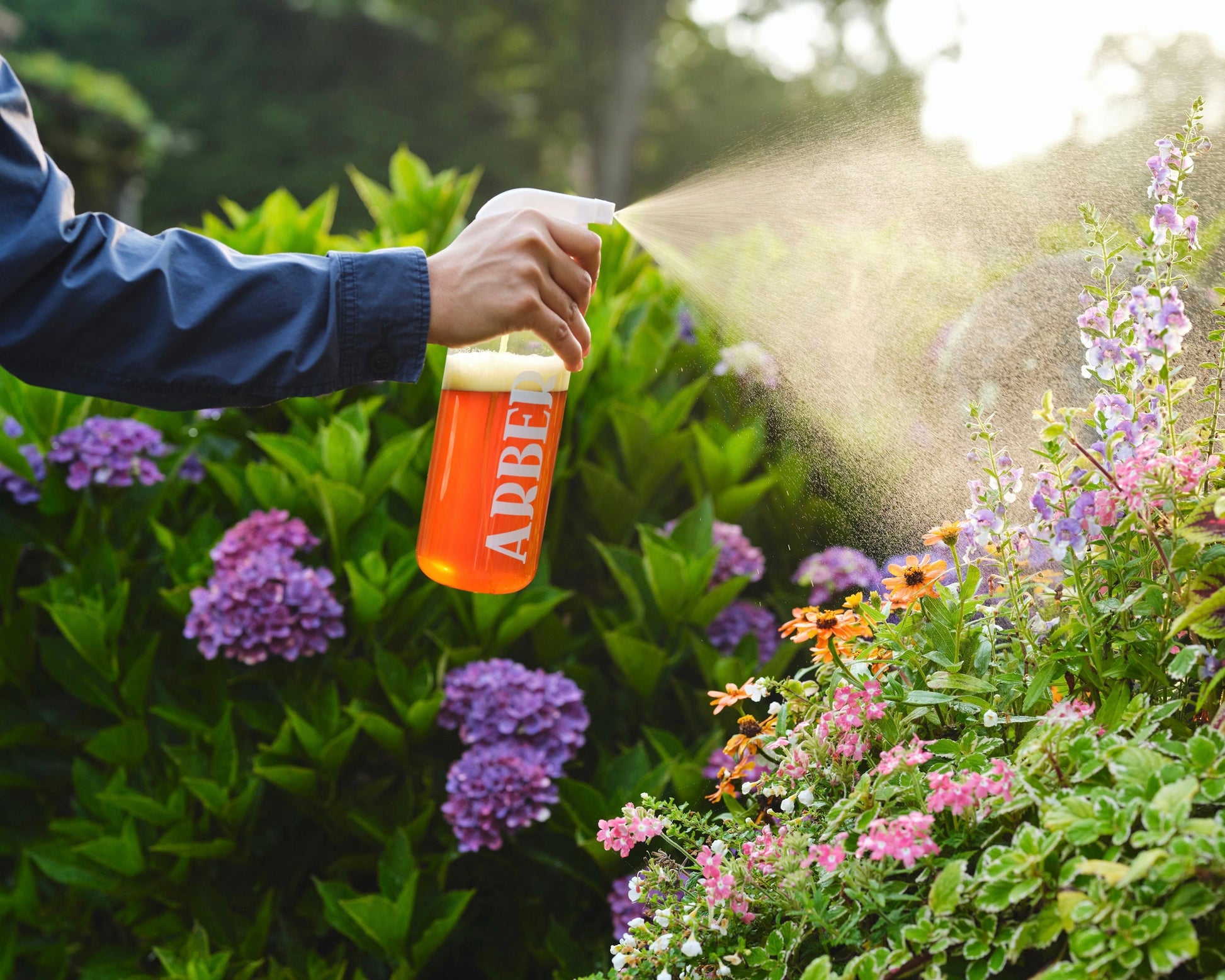Using an Arber Spray Bottle, a person mists blooming garden flowers, supporting plant health as sunlight catches the fine spray and bright blossoms in the background.