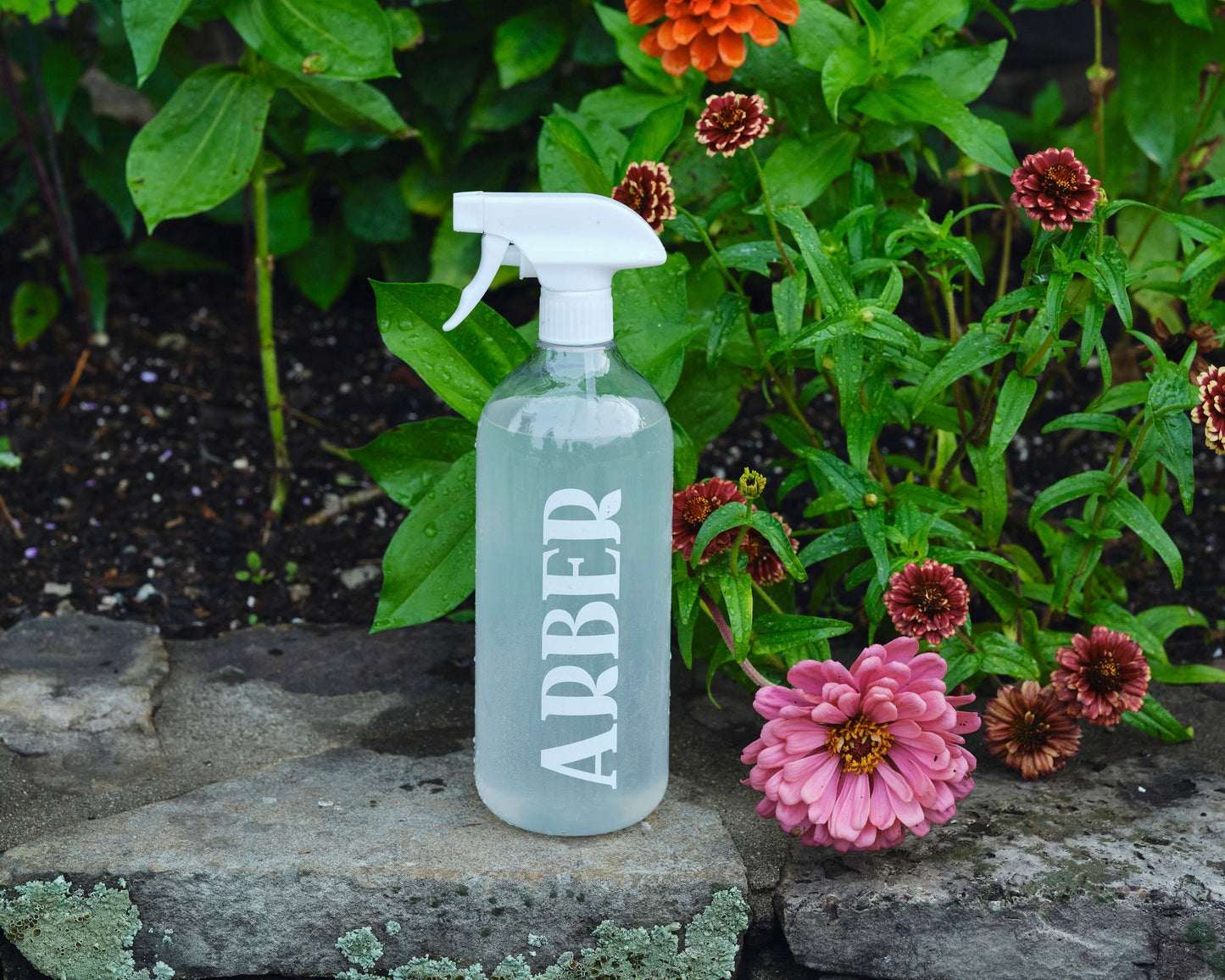 An Arber Spray Bottle sits on a stone ledge with blooming pink and red flowers, highlighting plant wellness and garden hydration among lush green foliage.