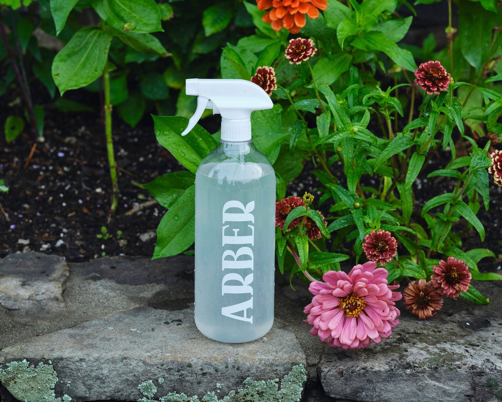 An Arber Spray Bottle sits on a stone ledge with blooming pink and red flowers, highlighting plant wellness and garden hydration among lush green foliage.