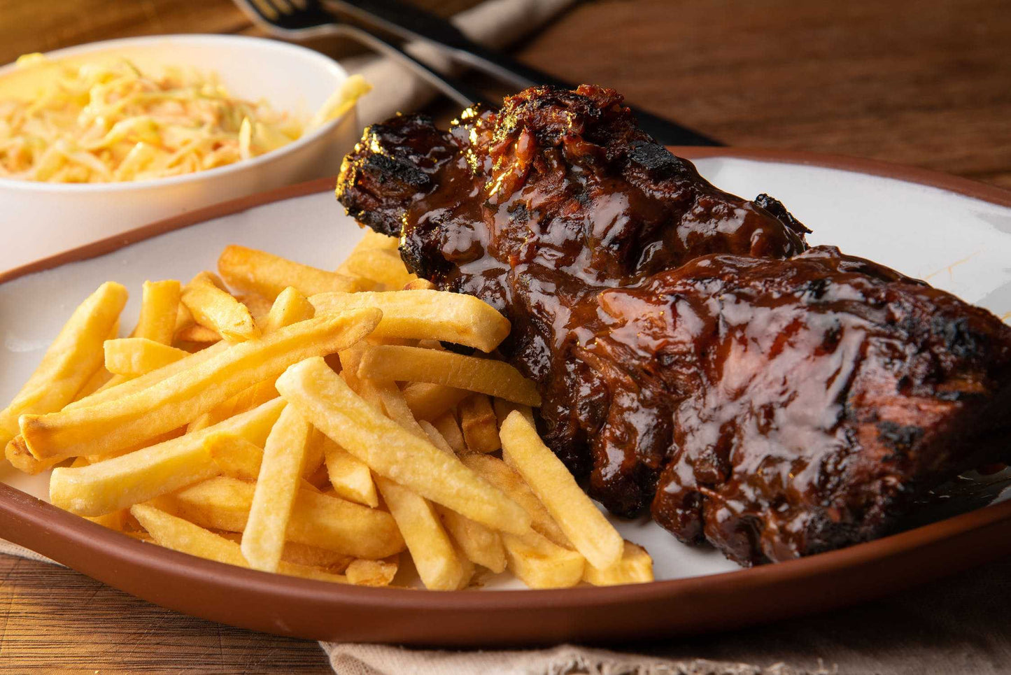 A plate of glazed barbecue ribs brushed with Weeks Honey Farms Raw Buckwheat Honey, served with golden fries and coleslaw in a white bowl on a wooden table.