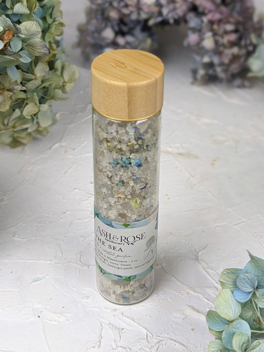 A clear glass BY THE SEA Bath Soak Vial by Ash & Rose, filled with colorful sea salt, sits on a textured white surface surrounded by dried hydrangea flowers. The vial features a wooden lid and an Ash & Rose label.