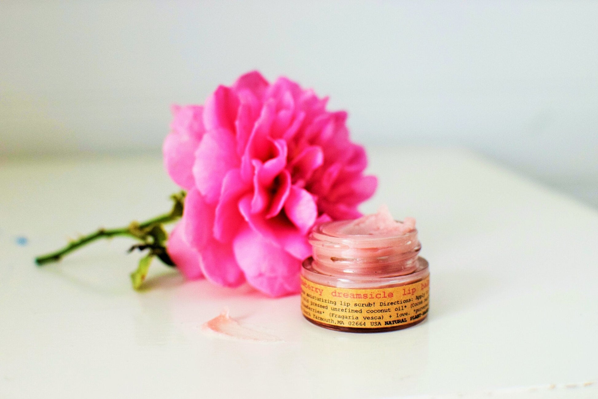 Plant Makeup Strawberry Dreamsicle Lip Balm Scrub in a clear jar with a pink and white creamy scrub topped with a fresh strawberry.