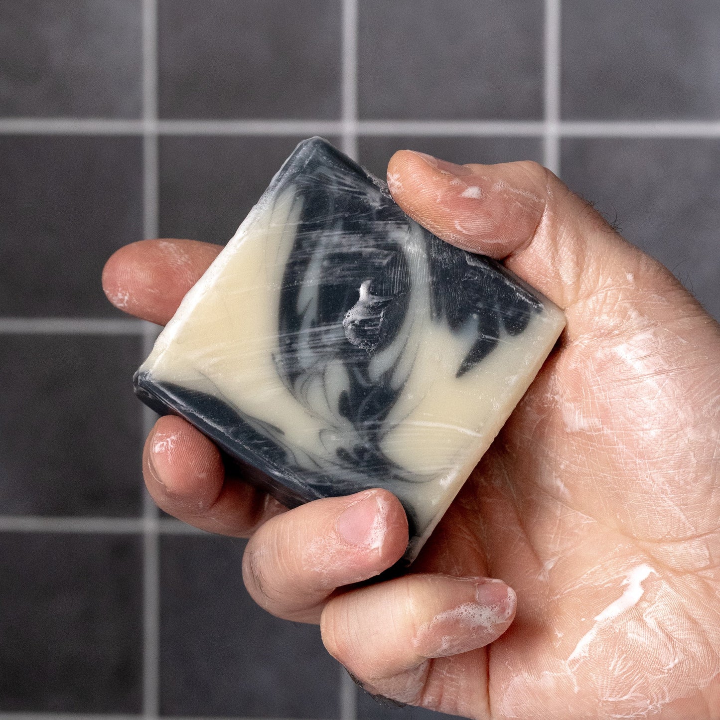 A hand holds Rustic Strength’s Bar Soap | Detoxifying Charcoal, featuring black and white marbling and visible lather, against a tiled background—ideal for sensitive skin.