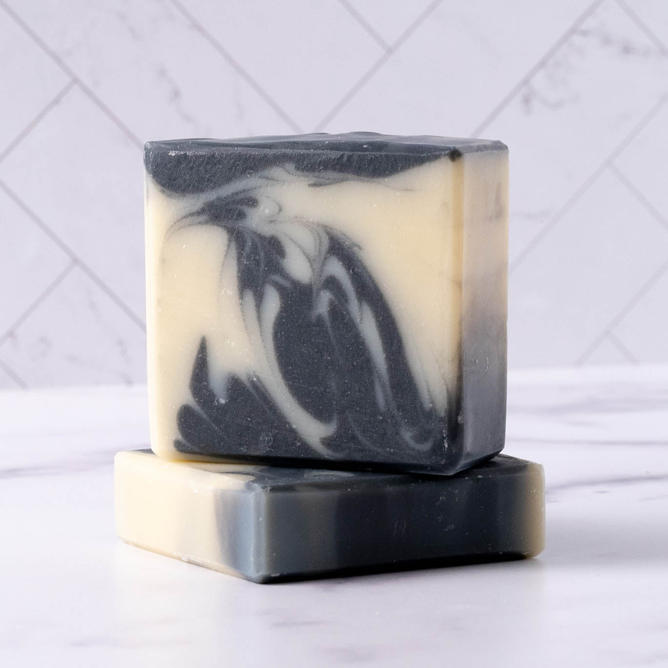 Two square Rustic Strength Bar Soap | Detoxifying Charcoal bars, stacked upright, display a marbled black and cream pattern. Designed for sensitive skin, they sit against white geometric tiles.