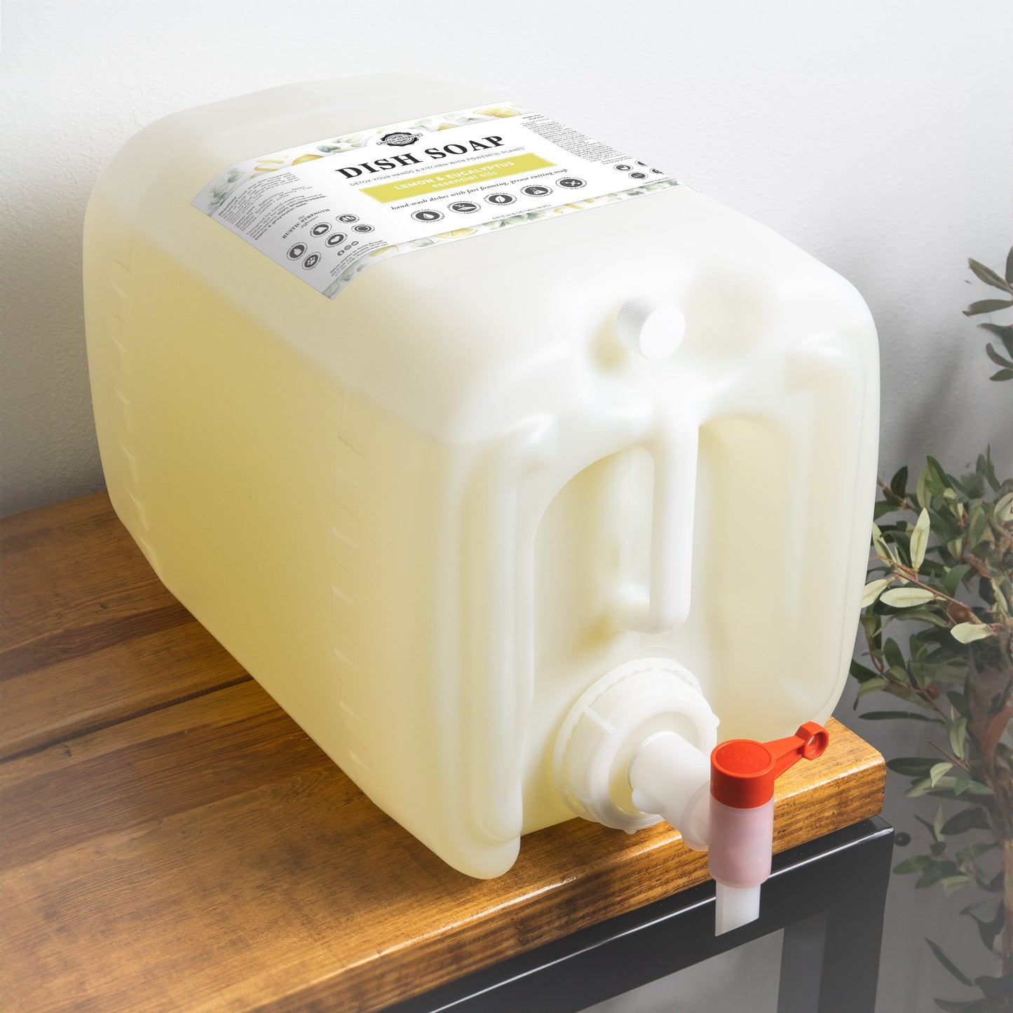 A large, translucent container of Rustic Strength Dish Soap | Lemon & Eucalyptus with a red-and-white spigot and branded label sits on a wooden surface near a leafy plant.