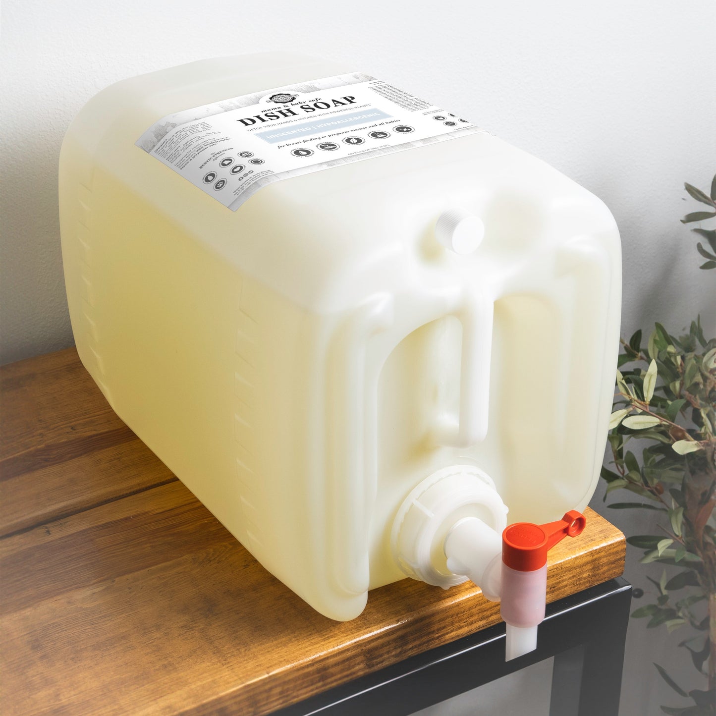 A large white plastic container of Rustic Strength Dish Soap | Mama + Baby Safe, filled with yellowish unscented liquid and equipped with a spigot, sits on a wooden surface beside a green plant.