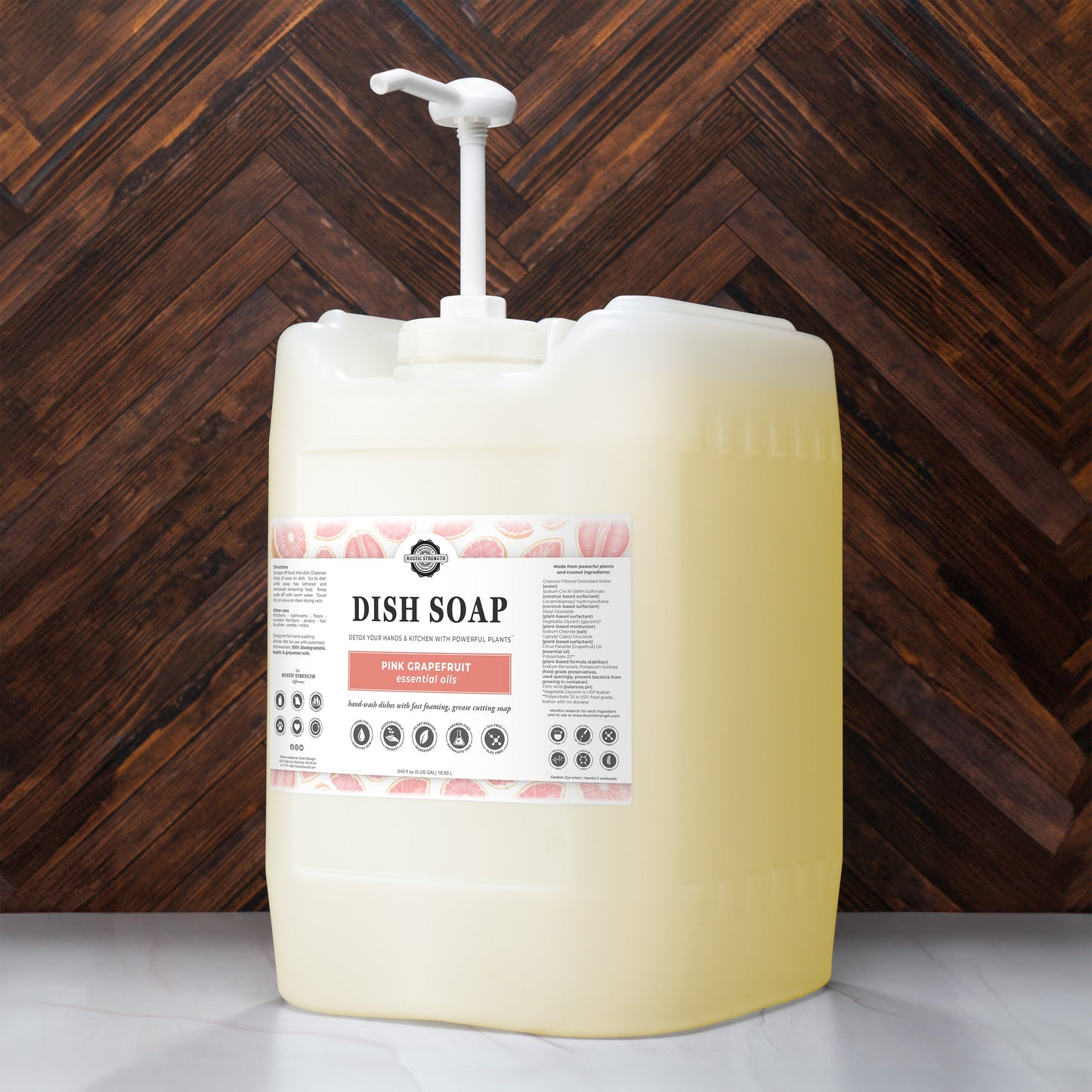 A large, translucent plastic container of Rustic Strength Dish Soap | Pink Grapefruit with a pump dispenser sits on a wooden surface. The label highlights its fresh grapefruit scent against the dark wood background.
