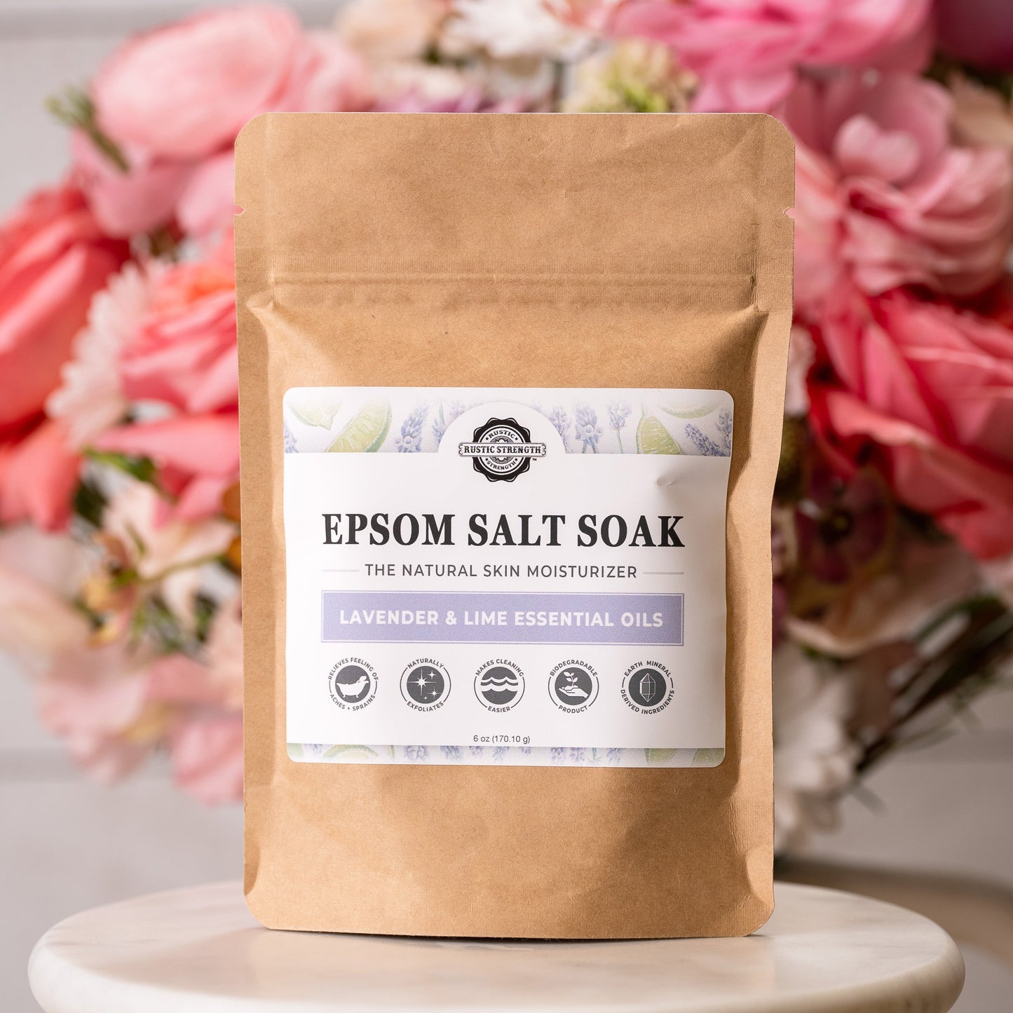A brown paper bag labeled Rustic Strength Epsom Salt Soak | Lavender & Lime stands before blurred pink and red flowers. The non-toxic label displays product icons and key information.