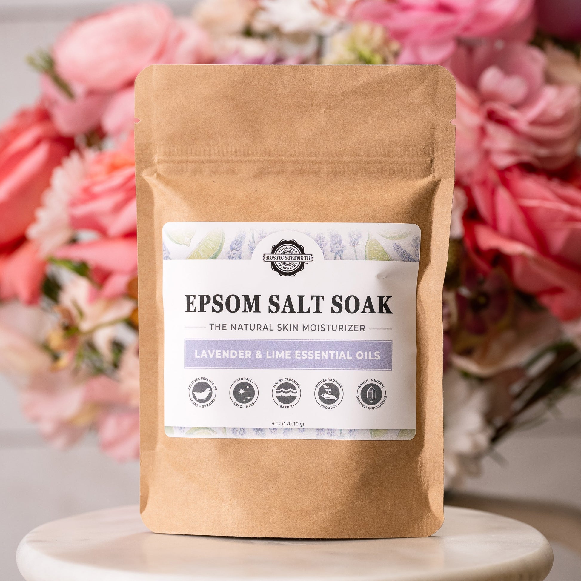 A brown paper bag labeled Rustic Strength Epsom Salt Soak | Lavender & Lime stands before blurred pink and red flowers. The non-toxic label displays product icons and key information.