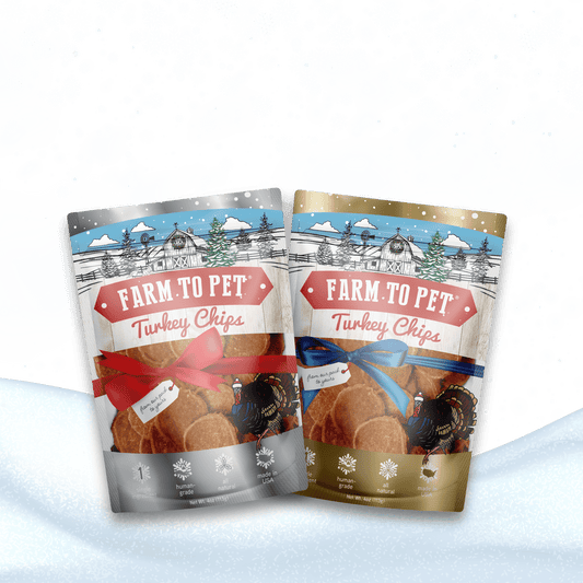 Two Farm To Pet Holiday Chips for Dogs bags, ideal low-allergen training treats, sit in snow—one silver with a red ribbon and one gold with a blue ribbon—set against a green, white, snowflake-filled background.