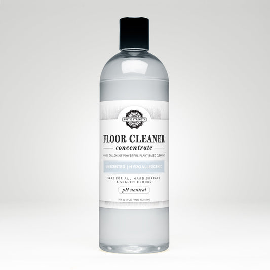 A clear plastic bottle labeled Rustic Strength Floor Cleaner Concentrate | Unscented, featuring hypoallergenic, pH neutral, and non-toxic qualities, contains 16.9 fl oz of floor cleaner liquid set against a plain white background.