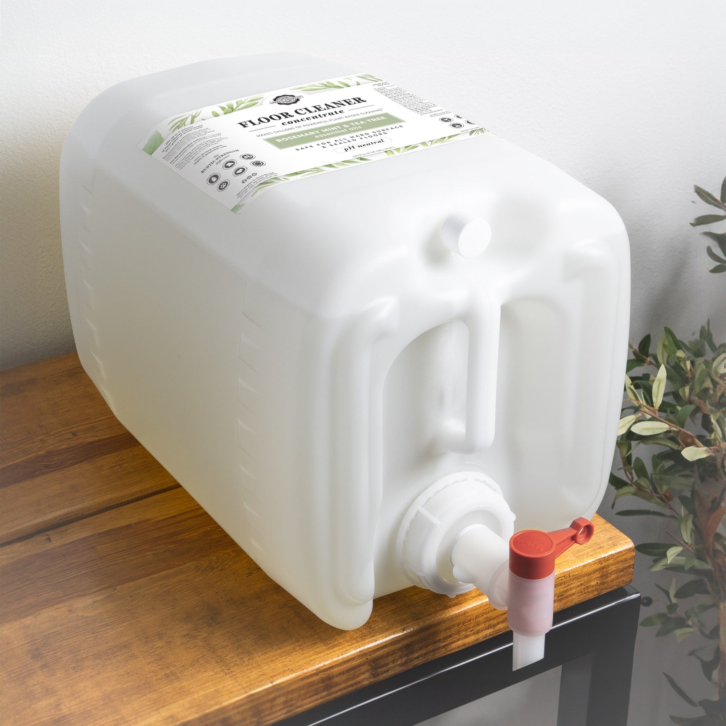 A white plastic container of Rustic Strength Floor Cleaner Concentrate | Rosemary, Mint & Tea Tree with a spout lies on its side on wood. The label displays text and green graphics; a leafy plant is partially visible behind it.