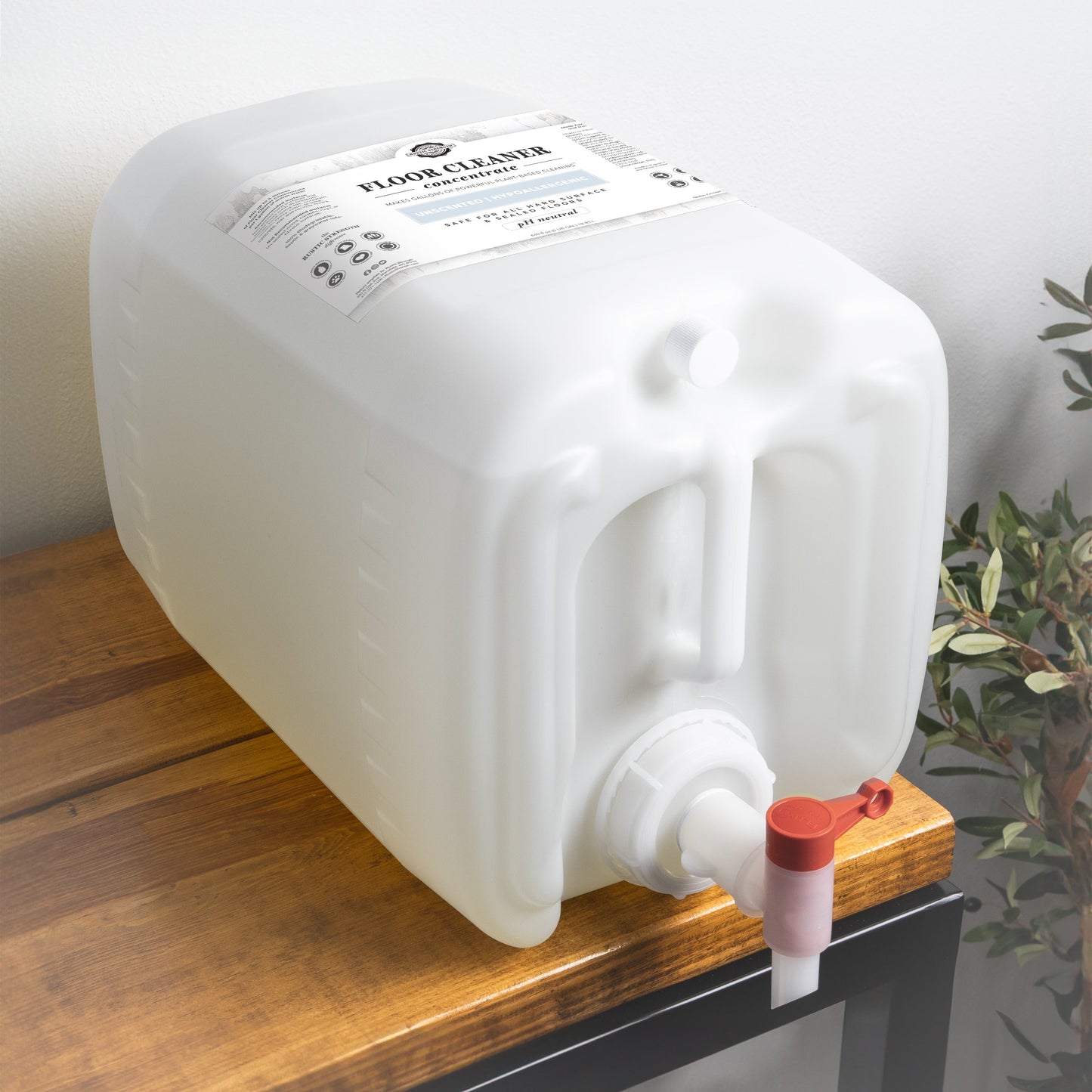 A large white plastic container of Rustic Strength Floor Cleaner Concentrate | Unscented, with a label and spigot for dispensing liquid, sits on a wooden table near a wall and plant. The non-toxic cleaner comes in an easy-to-use dispenser.