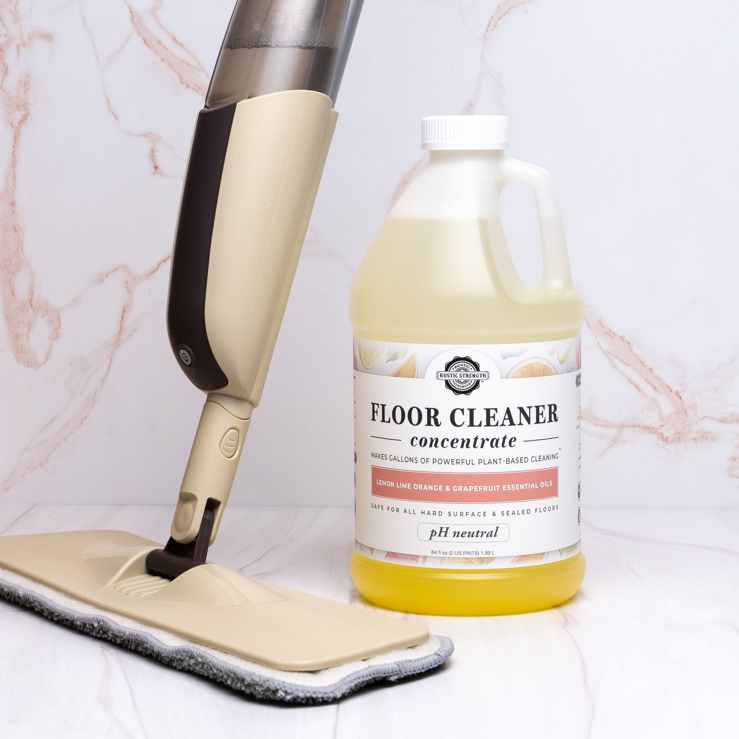 A beige mop leans against a large bottle of Rustic Strength Floor Cleaner Concentrate (Lemon, Lime, Orange & Grapefruit) filled with yellow liquid, set on a marble-patterned surface with a light pink and white background.