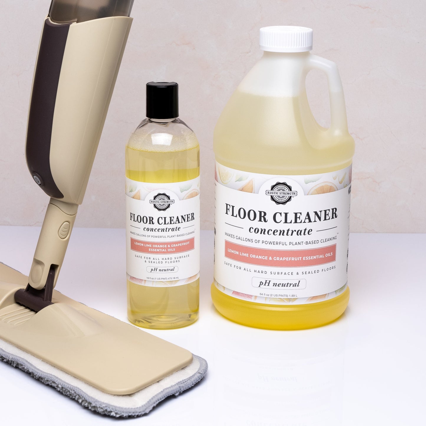 Two bottles (large and small) of Rustic Strength Floor Cleaner Concentrate | Lemon, Lime, Orange & Grapefruit sit beside a beige mop with a white pad. Labels highlight non-toxic cleaning powered by essential oils.