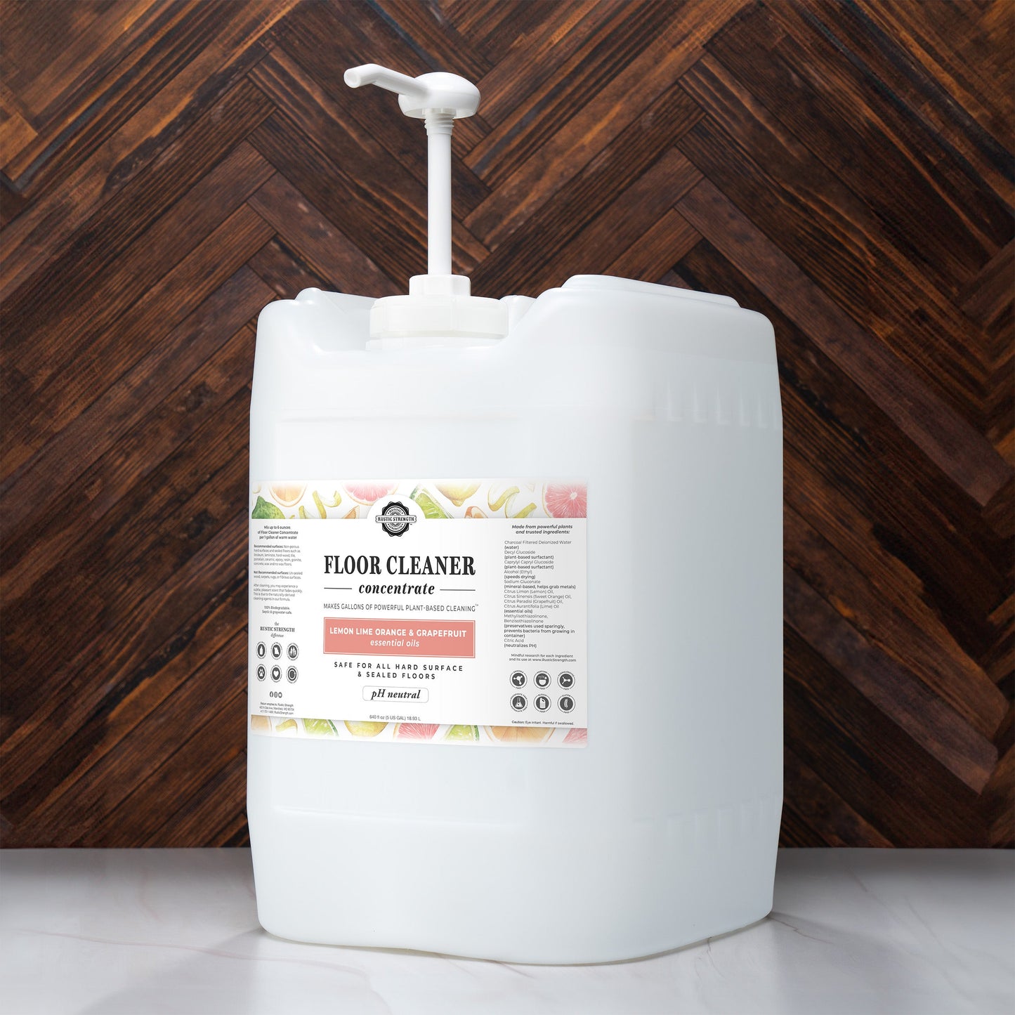 A large white Rustic Strength container with a pump dispenser, labeled Floor Cleaner Concentrate | Lemon, Lime, Orange & Grapefruit, sits on a herringbone-patterned wooden floor. The non-toxic label features product details and instructions.