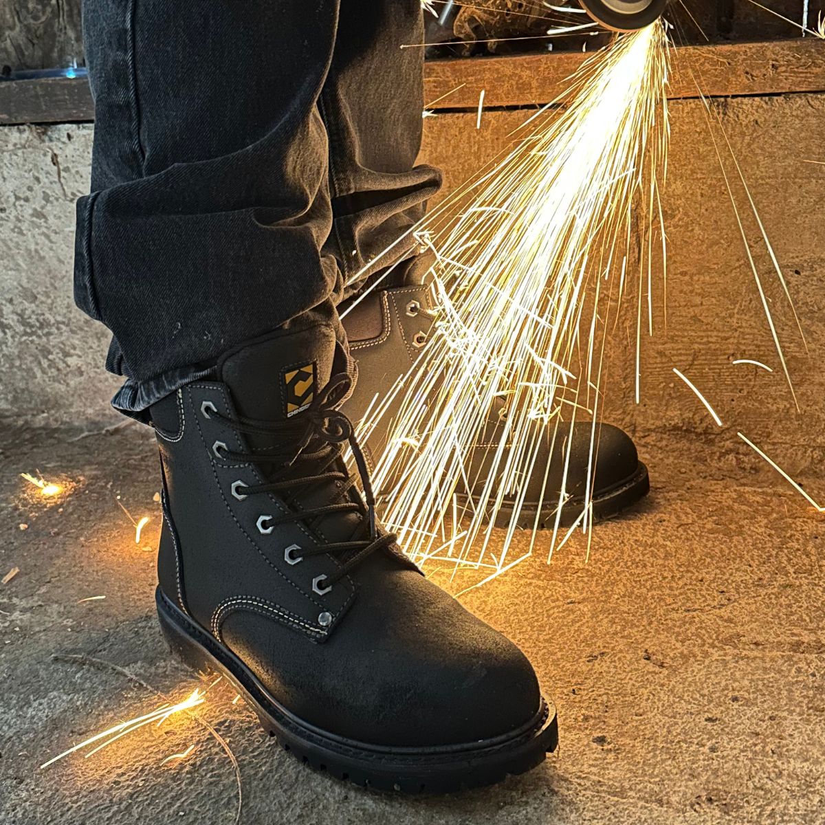 Wearing Chinook® Footwears Oil Rigger 6” Steel Toe Men’s Work Boot in black and dark jeans, a worker stands on concrete as bright sparks fly from a power tool nearby.