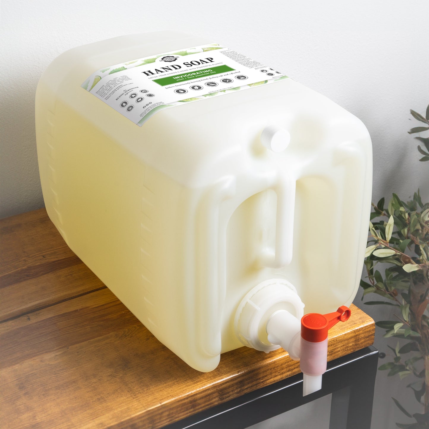 A large, rectangular Rustic Strength Hand Soap | Invigorating plastic container with a spout sits on a wooden table near a leafy plant. The light yellow container has a label on top.