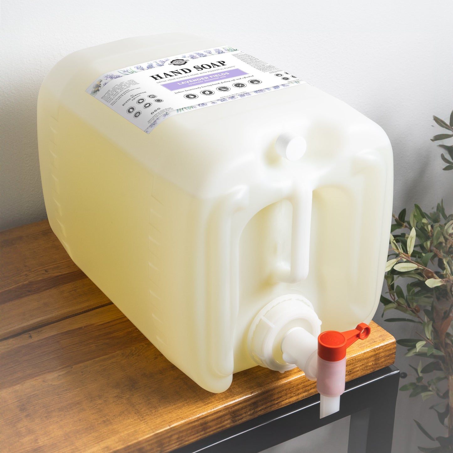 A large, translucent plastic container labeled Rustic Strength Hand Soap | Lavender Fields sits on a wooden surface. The white spout with a red lever dispenses light yellow biodegradable soap. A green plant is nearby.