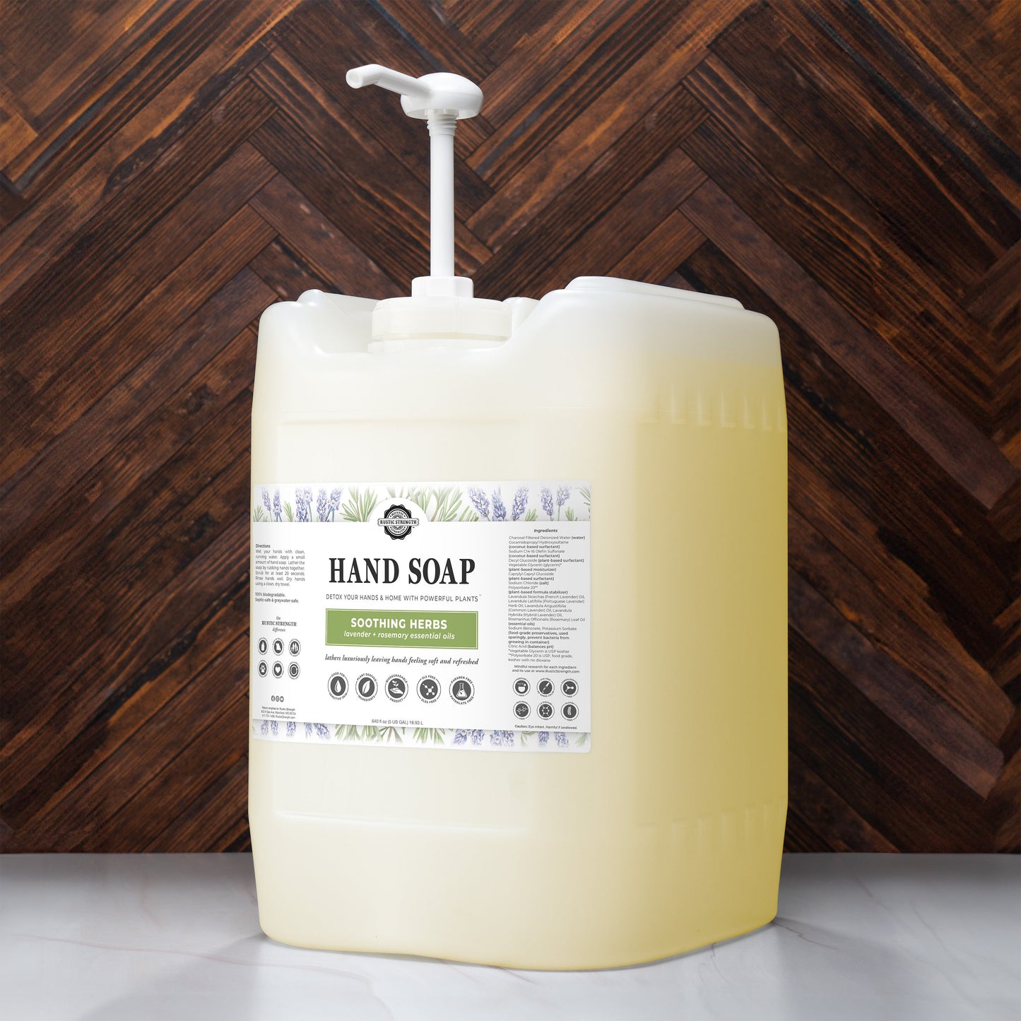 A large, translucent bottle of Rustic Strength Hand Soap | Soothing Herbs with a pump sits on a wooden surface. The label features botanical illustrations and product details.