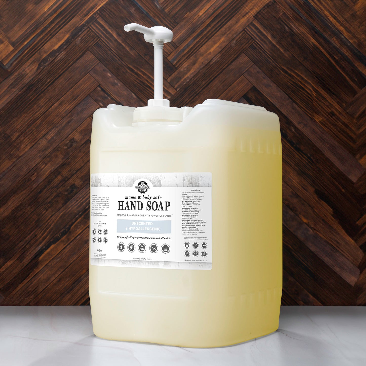 A large, translucent plastic container of Rustic Strength Hand Soap | Mama + Baby Safe with a pump sits on the countertop. The pale yellow soap’s label displays usage instructions and icons. A brown herringbone pattern is in the background.