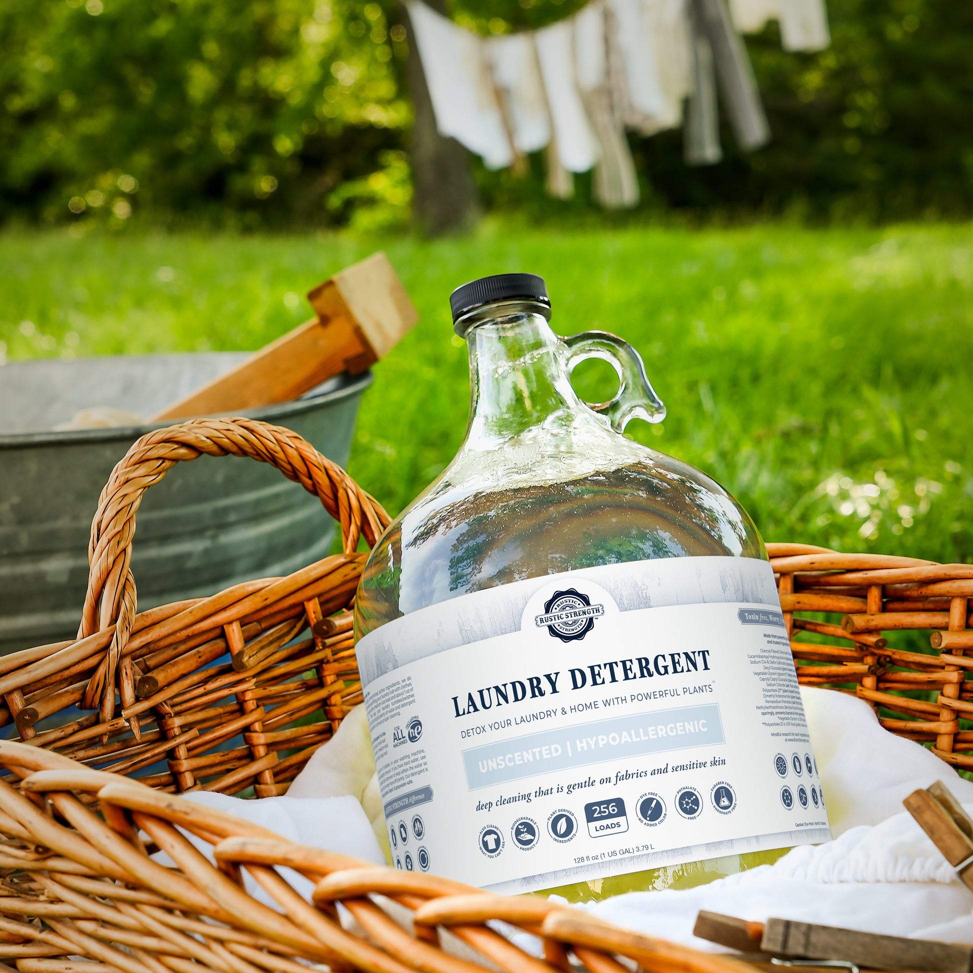 A large glass jug of Rustic Strength Laundry Detergent | Lavender, biodegradable and hypoallergenic, rests in a wicker basket outdoors beside a metal wash tub, white cloth, and laundry drying on a clothesline.