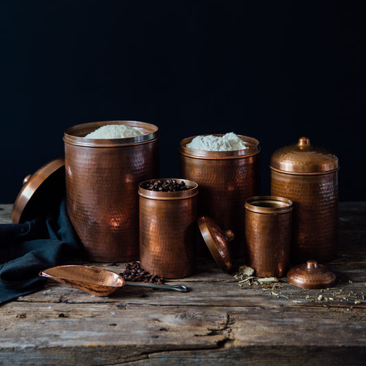 Set of Sertodo copper kitchen canisters in various sizes with polished, reflective surfaces and wooden lids, on a kitchen countertop.