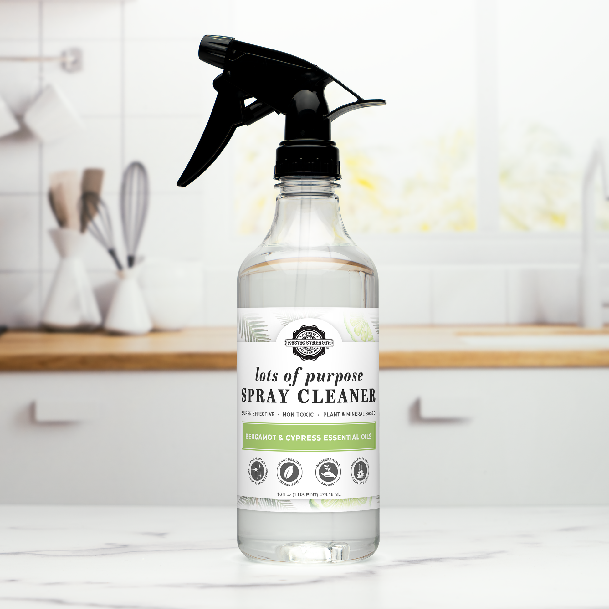 A clear bottle of Rustic Strength Lots of Purpose Spray Cleaner | Bergamot & Cypress sits on a white kitchen counter, with a modern kitchen background and utensils. The plant-based formula features bergamot and cypress essential oils.