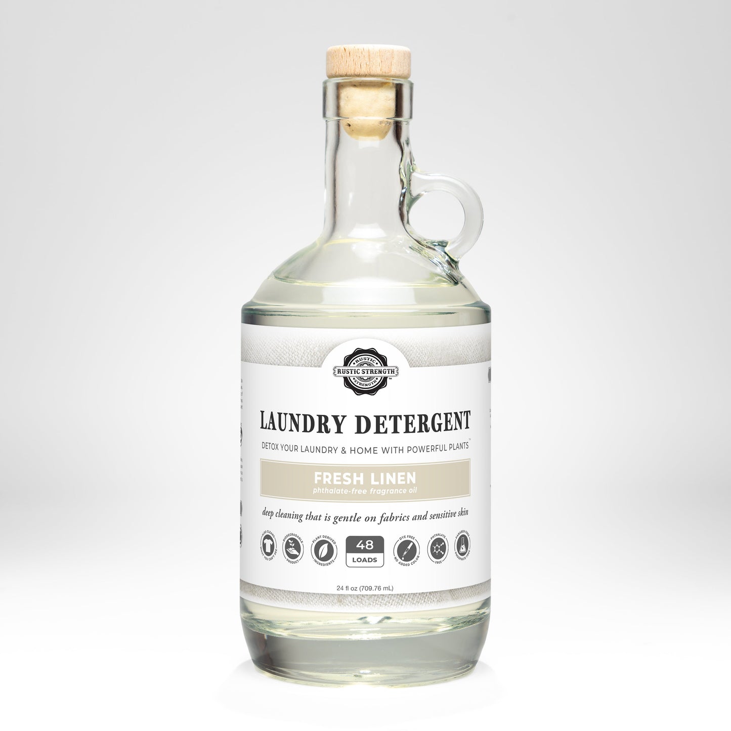 A clear glass bottle with a wooden cork holds Rustic Strength Laundry Detergent | Fresh Linen, 24 fl oz (48 loads). The label highlights plant-based, hypoallergenic cleaning with icons for gentle, biodegradable detergent.
