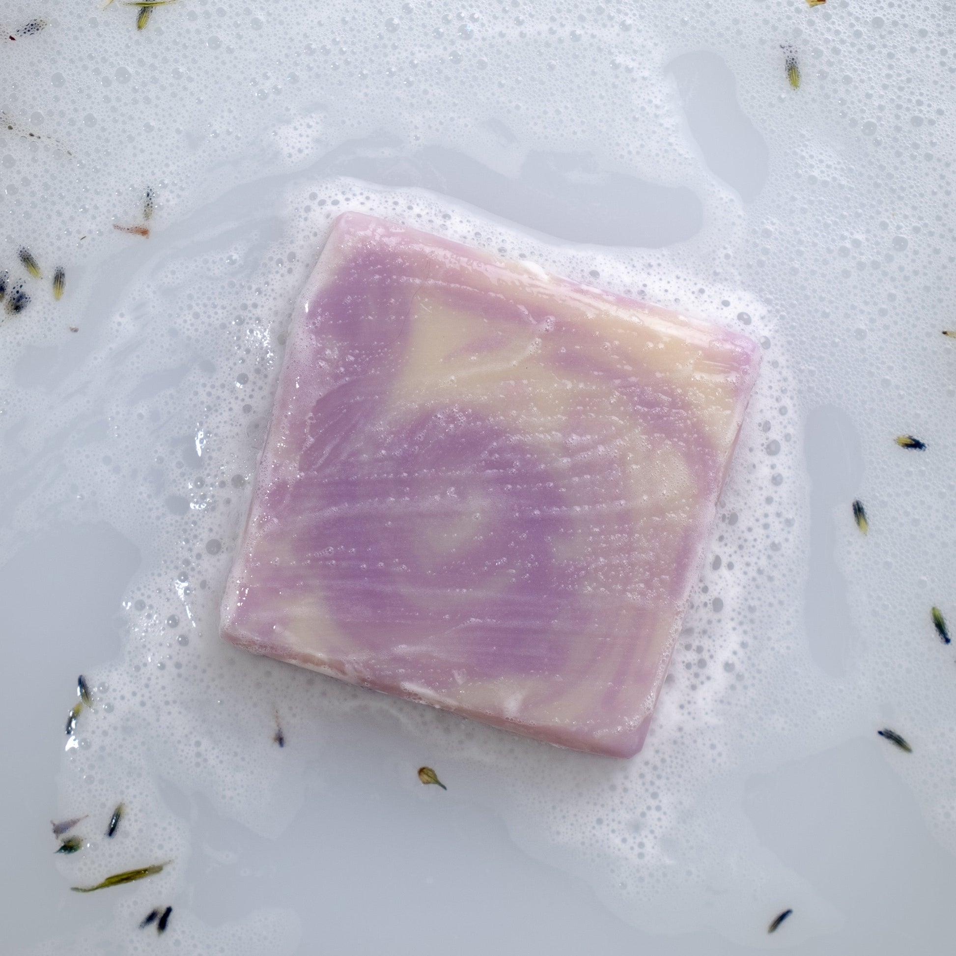 A square bar of Rustic Strength Bar Soap | Lavender, with purple and white marbling, rests in soapy water among bubbles and lavender sprigs—ideal for sensitive skin.
