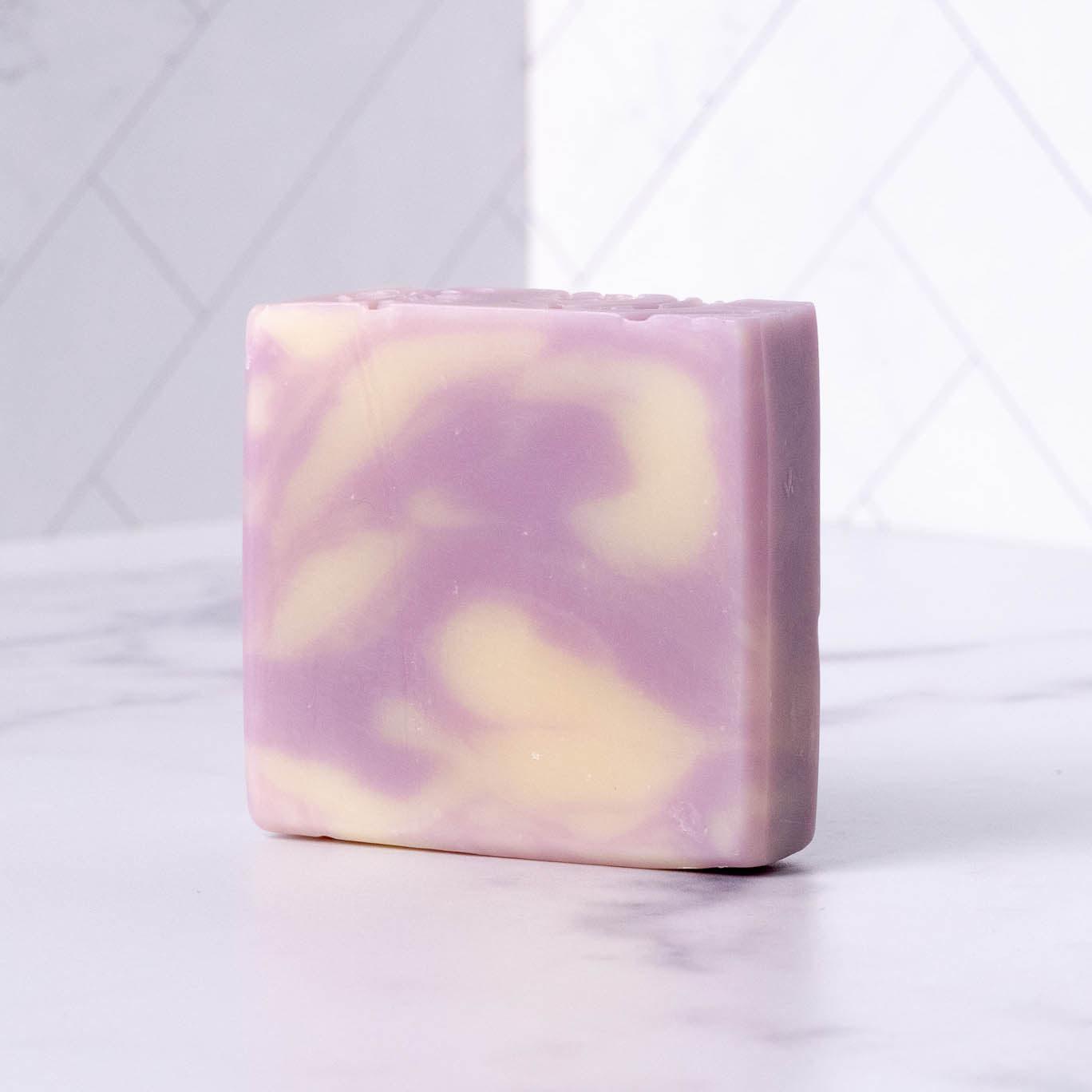 Rustic Strengths Bar Soap | Lavender, featuring a swirled pattern of light purple and cream, is perfect for sensitive skin and sits beautifully on a white marble surface with a tiled background.