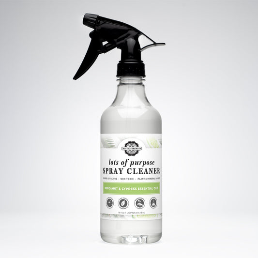 A clear spray bottle labeled Rustic Strength Lots of Purpose Spray Cleaner | Bergamot & Cypress has a black trigger nozzle with green accents and icons showcasing its plant-based, non-toxic multi-surface formula, set against a white background.