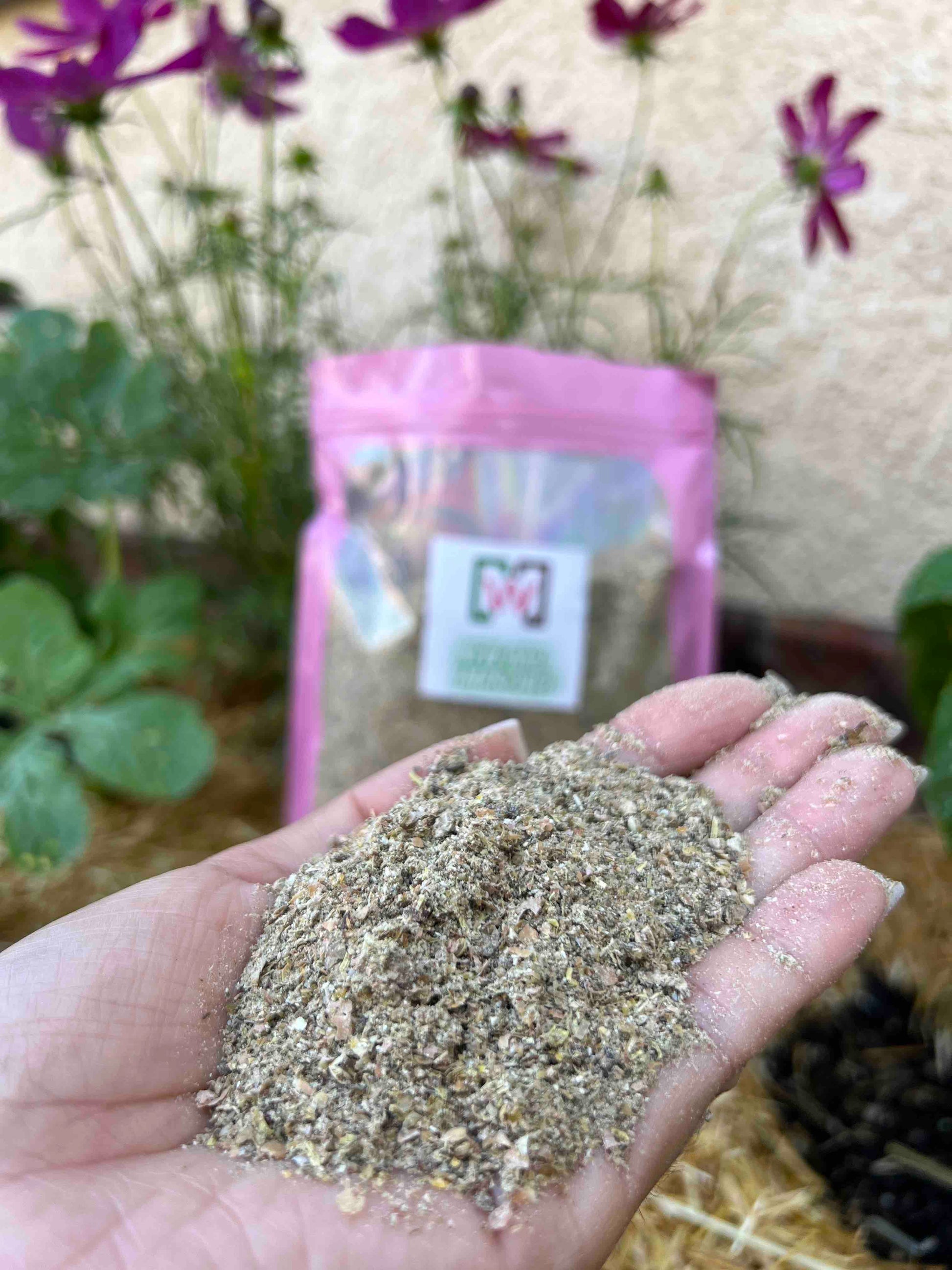 A hand holds a pile of coarse, brownish Memes Premium Worm Chow in front of a pink resealable Memes Worms bag, with flowering plants blurred in the background—perfect for your composting bin.