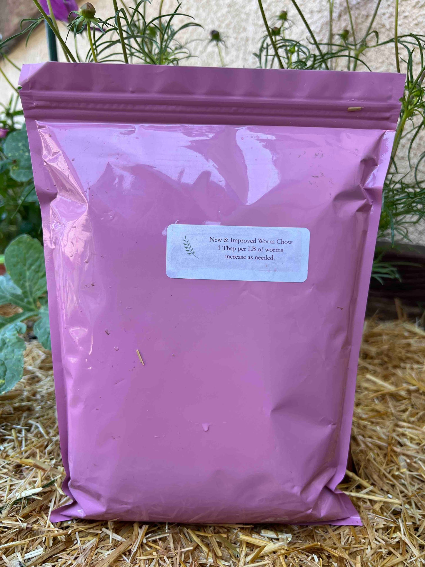A pink resealable bag labeled Memes Premium Worm Chow by Memes Worms rests on straw with green plants behind it. The label includes instructions for feeding worms in your composting bin.