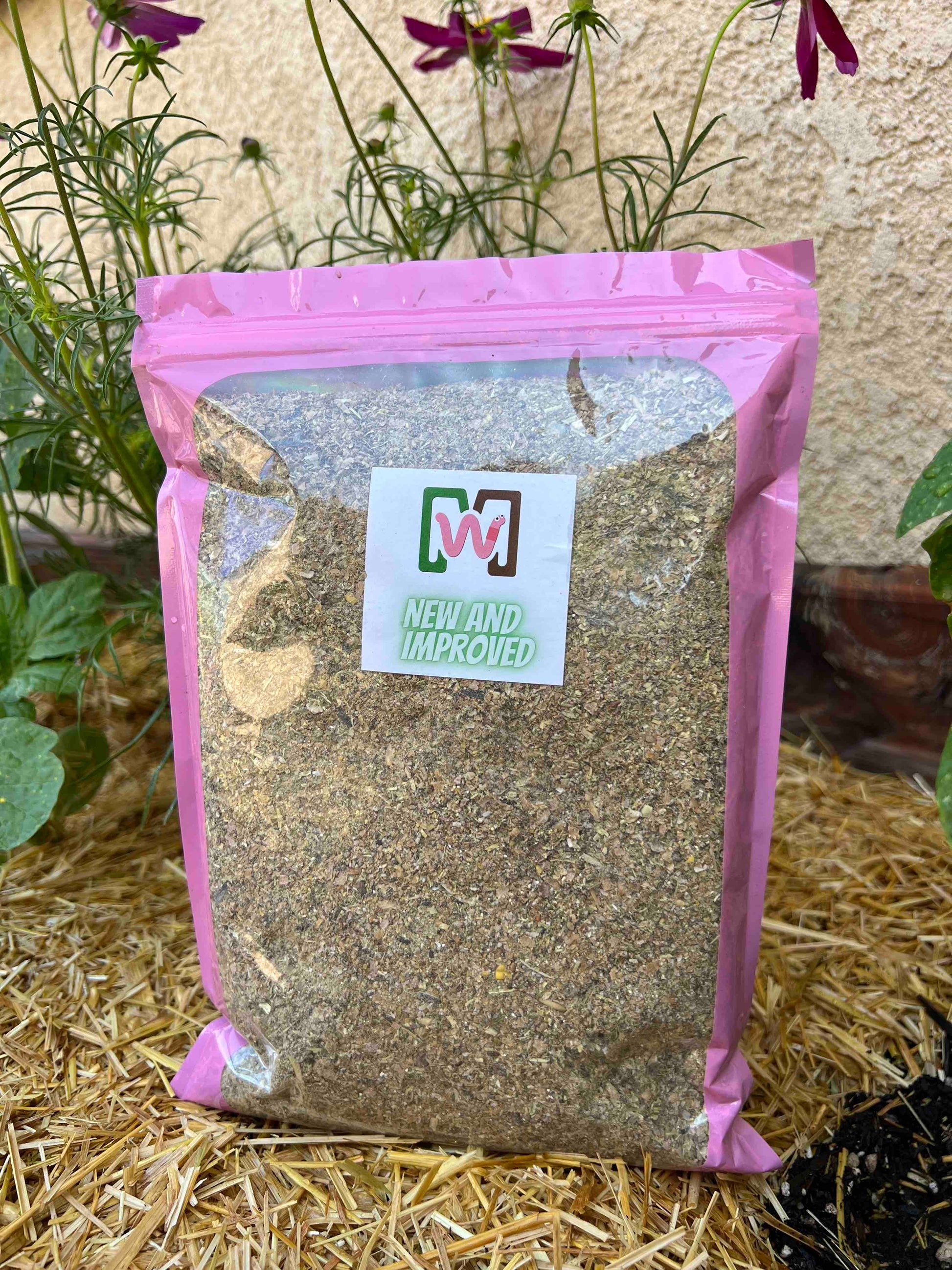 A clear plastic bag with a pink border holds Memes Premium Worm Chow by Memes Worms. The bag features a white label with NEW AND IMPROVED in green and is shown outdoors among plants, perfect for your composting bin.