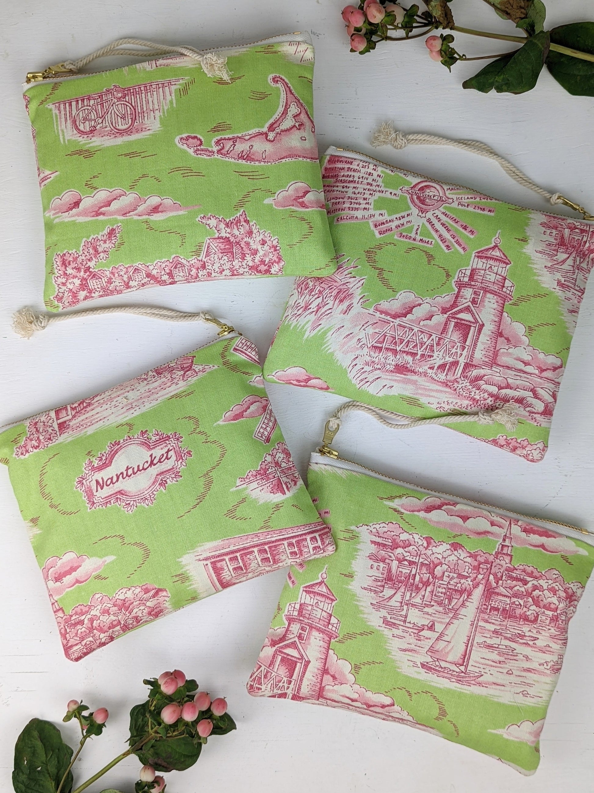 Four Nantucket Purses by Ash & Rose—handmade in Massachusetts with green and pink fabric, gold zippers, and Nantucket-inspired designs—are displayed on a white surface with pink berries and green leaves.