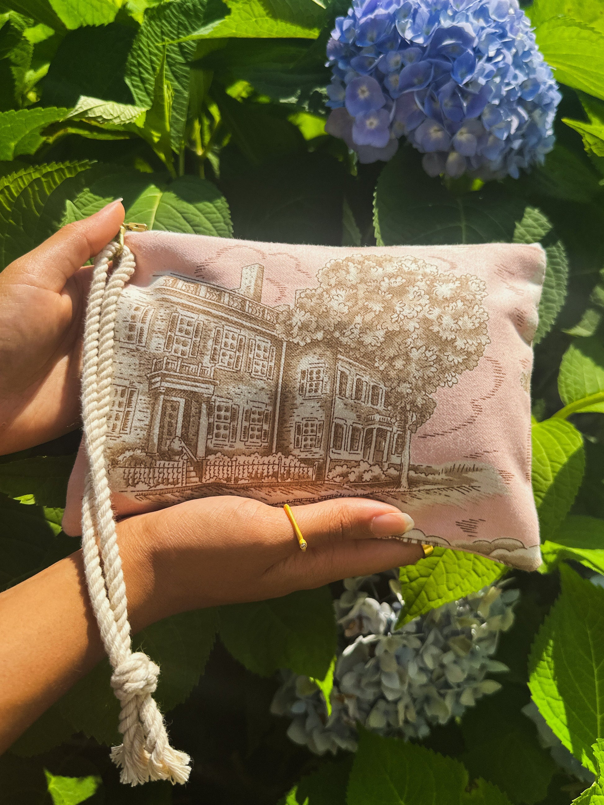 A person holds the Nantucket Purse by Ash & Rose, a limited edition handmade pouch from Massachusetts featuring a house and trees print with a white rope handle. Lush green leaves and blooming blue hydrangeas appear in the background.