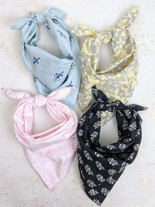 Four Ash & Rose Organic Cotton Bandanas in light blue, yellow, pink, and black, each tied in a loose knot, are arranged in a grid on a white surface with gold speckles. All bandanas are fair trade and feature different patterns.