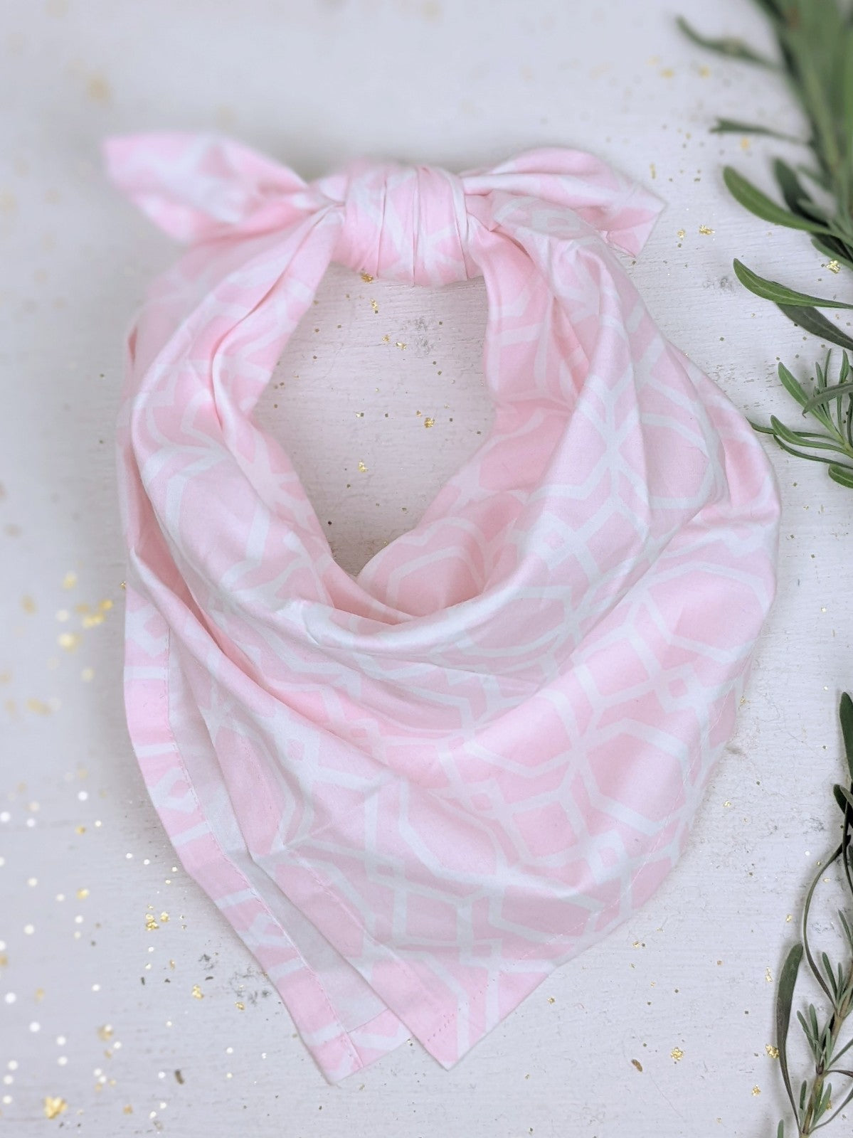 The Ash & Rose Organic Cotton Bandana in soft pink with a subtle geometric pattern is tied at the top and laid flat on a light surface, surrounded by gold confetti and green foliage.