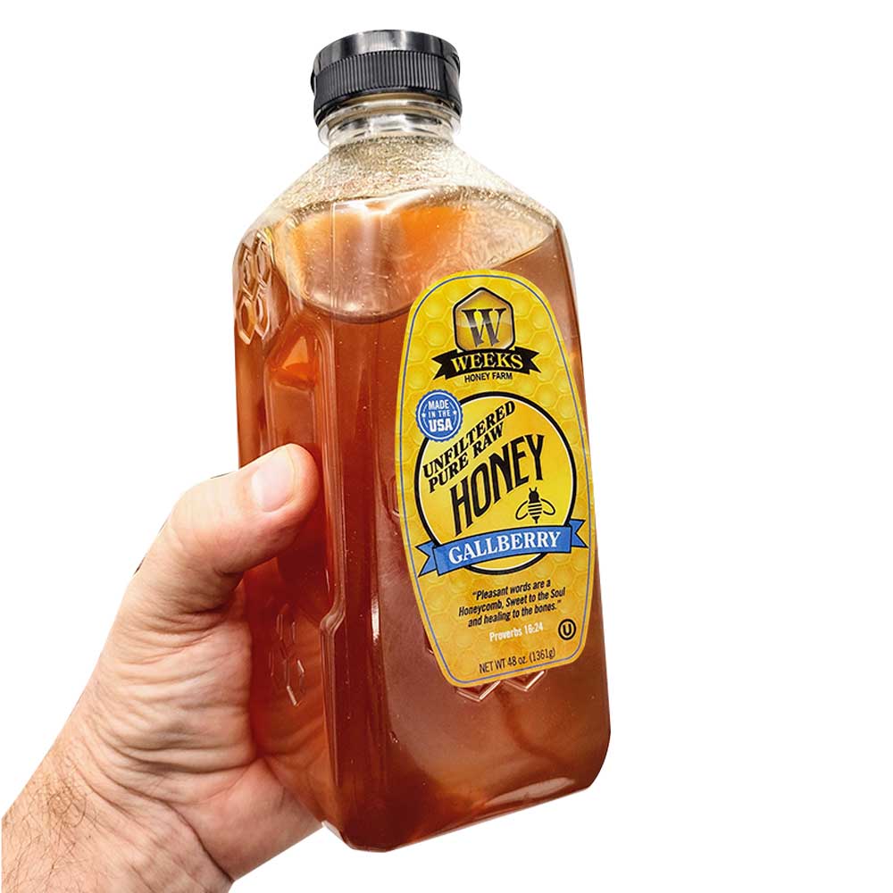 A hand holds a clear plastic bottle of Our Best All-Natural Pure Raw Gallberry Honey by Weeks Naturals | Weeks Honey Farm, featuring a yellow label and black cap, against a white background.