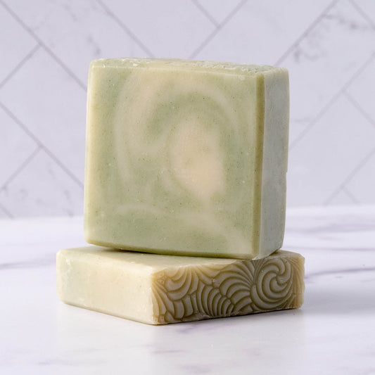 Two greenish-beige Rustic Strength Bar Soap | Peppermint Tea Tree bars sit on a white surface, one upright with a subtle swirl and the other flat showing texture, set against a marble tile background.