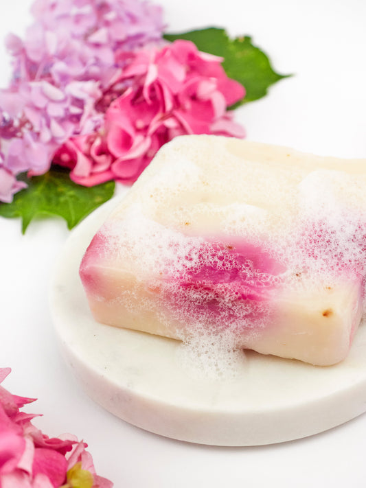 The Ash & Rose Pink Hydrangea Soap Scrub Bar, a pink and white handmade square soap, rests on a round marble dish with pink and purple hydrangeas, set against a white background.