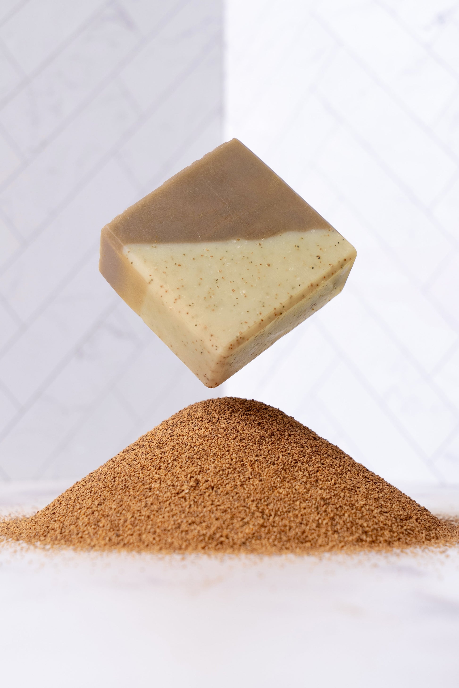 A beige and brown Rustic Strength Bar Soap | Sandalwood Patchouli, exfoliating with walnut, floats above a small heap of brown powder on a white surface against a white tiled background.